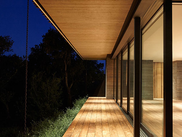 home-security Modern wooden deck at night, illuminated by warm recessed lighting from the overhanging roof, highlighting the sleek glass walls and blending seamlessly with the surrounding trees.