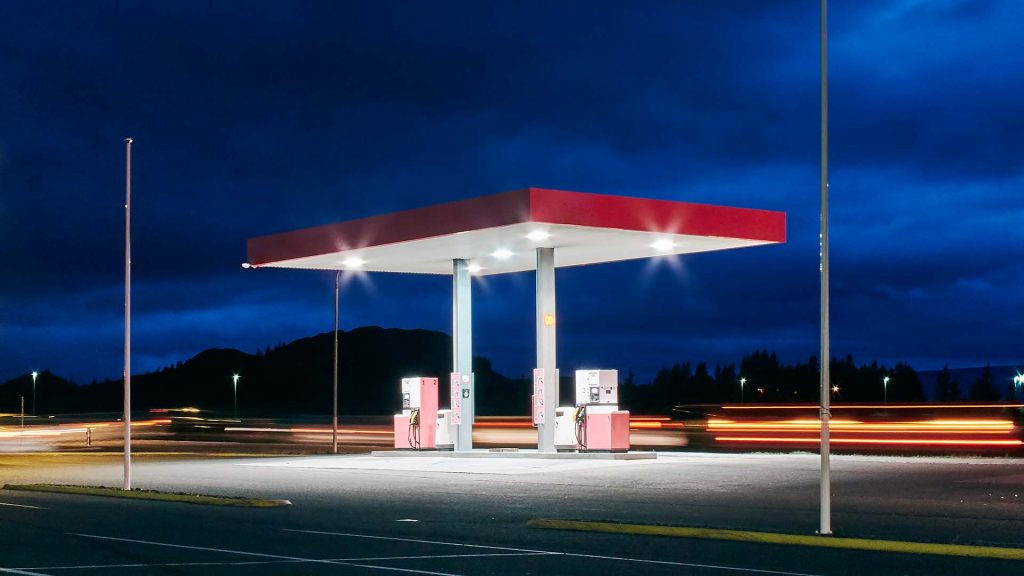 Gas station with a red canopy illuminated at night, surrounded by a dark sky and blurred streaks of car lights passing by, creating a dynamic and modern scene.