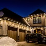 Exterior of a luxurious two-story house at night, adorned with warm, bright string lights along the roofline, illuminating the snow-covered landscape and enhancing the festive ambiance.