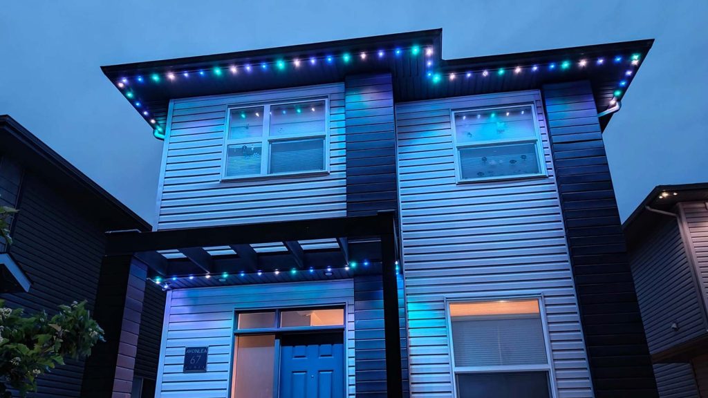 Two-story modern home at dusk with cool-toned LED puck lights installed along the roof eaves and porch overhang, casting a soft blue and green glow across the white vinyl siding and framing the blue front door.