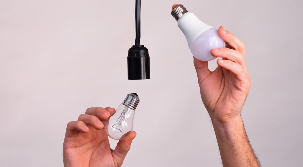 Two hands holding light bulbs under a hanging black pendant socket—one hand offers a clear incandescent bulb, the other holds a modern white-and-lavender LED bulb, illustrating swapping old technology for energy-efficient lighting.
