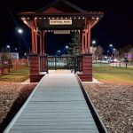 Nighttime view of a Central Park gazebo—red wooden structure with a ramped boardwalk leading up to it, outlined by a string of warm LED lights under the roof eaves, with surrounding park lights glowing in the background.