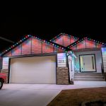 Nighttime front view of a single-story home (number 215) with multi-colour LED puck lights permanently mounted along the roofline eaves and gable peaks, casting vibrant accent lighting across the siding and shingle panels.