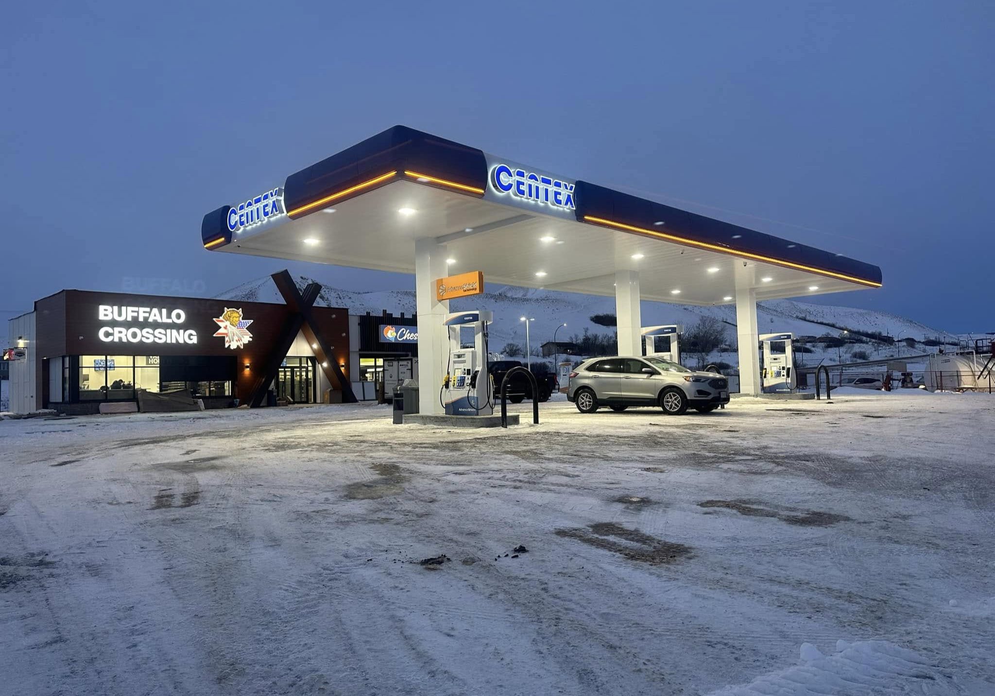 Nighttime view of a Centex gas station under a well-lit canopy—bright white LED fixtures recessed into the ceiling illuminate the pump area, while the adjacent Buffalo Crossing convenience store glows against a snowy foreground.